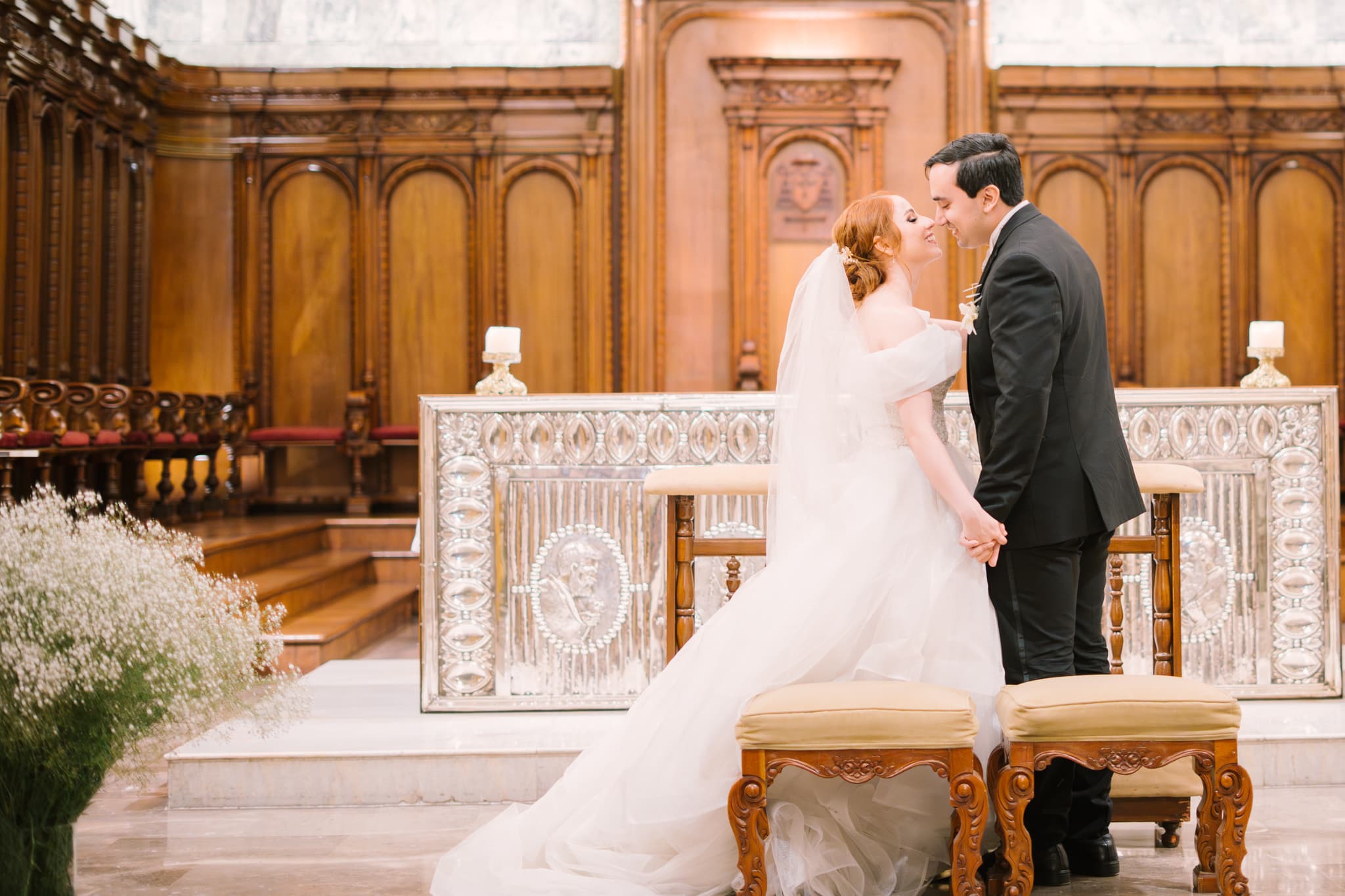 Novios tomados de la mano dándose un beso en el altar durante la ceremonia religiosa, con la novia en vestido blanco y el novio en traje negro.