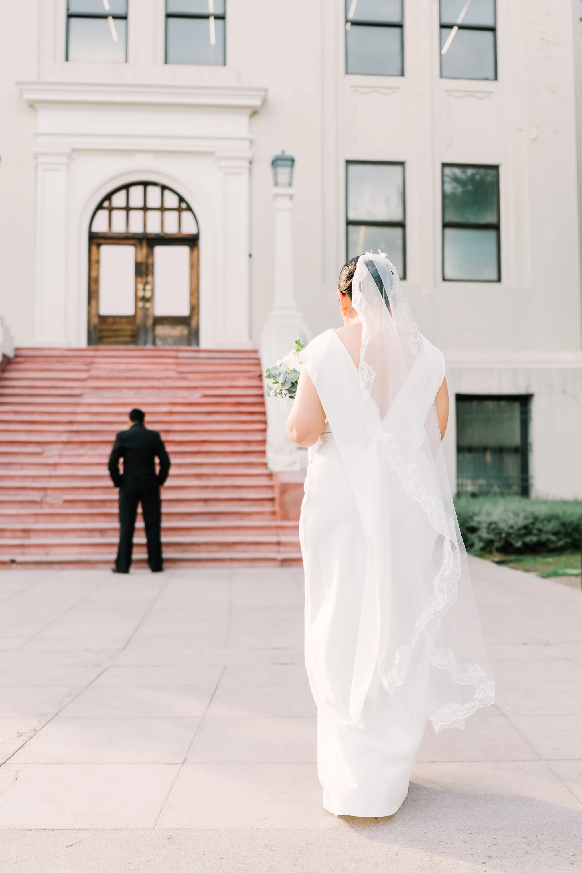 Vista trasera de la novia con vestido blanco de espalda en V y velo de encaje largo, caminando hacia el novio que está de espaldas frente a las escalinatas de piedra rosa del LABNL. La escena captura el momento previo al 'first look' frente a la fachada neoclásica del edificio.