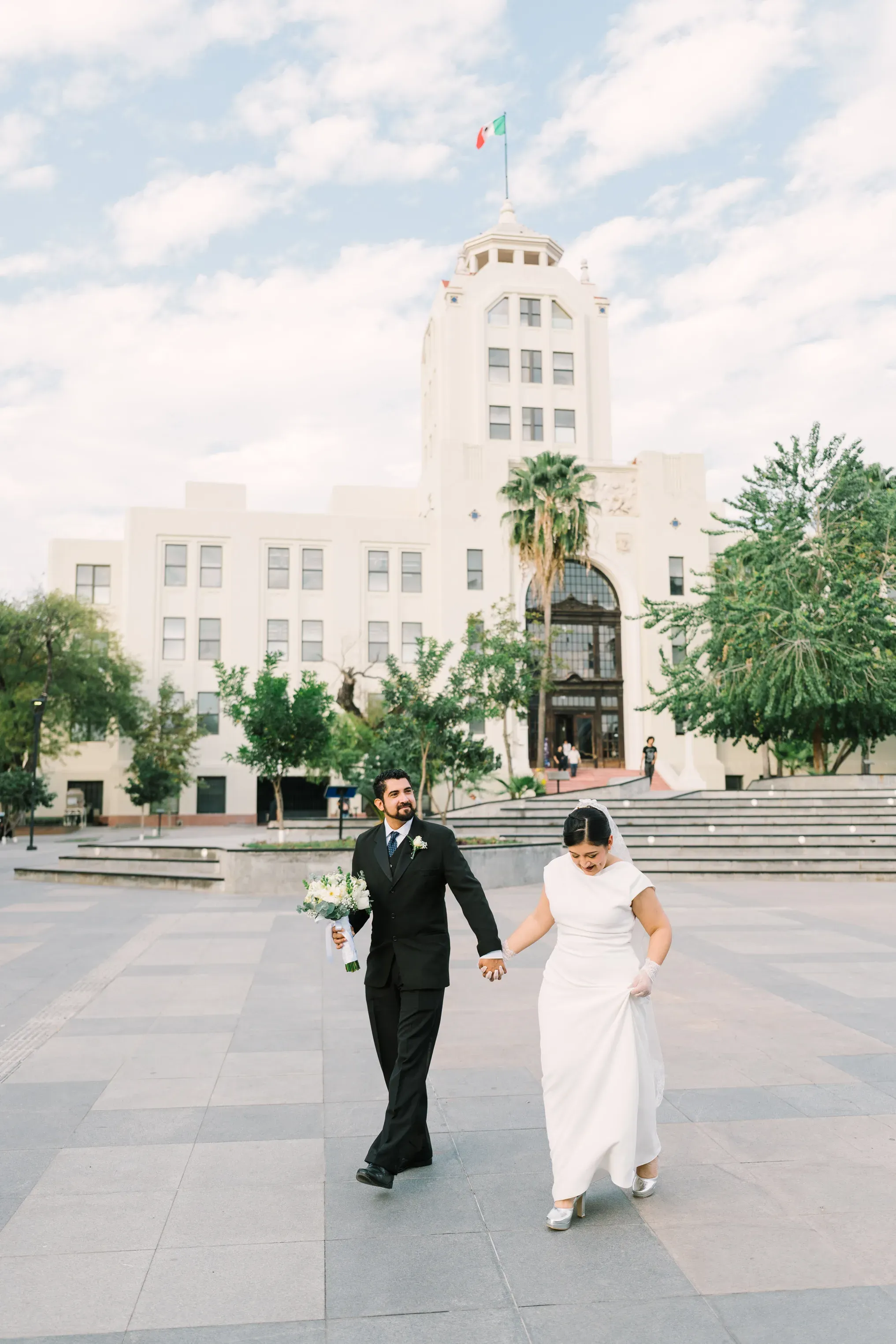 Amy y Jorge caminando tomados de la mano frente al Palacio Federal de Monterrey con la bandera mexicana al fondo