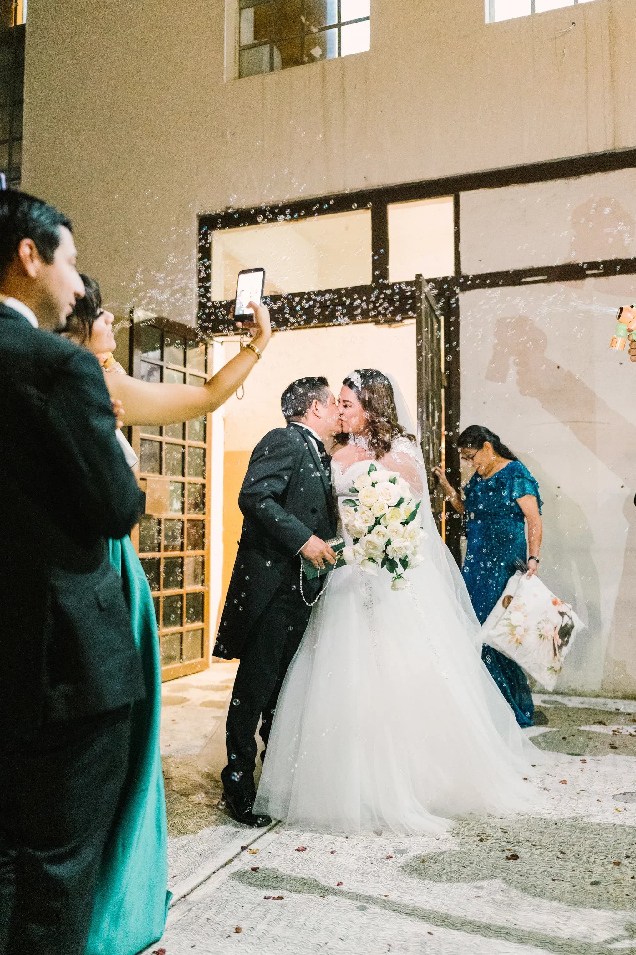 Novios besándose a la salida de la iglesia rodeados de burbujas y familiares aplaudiendo