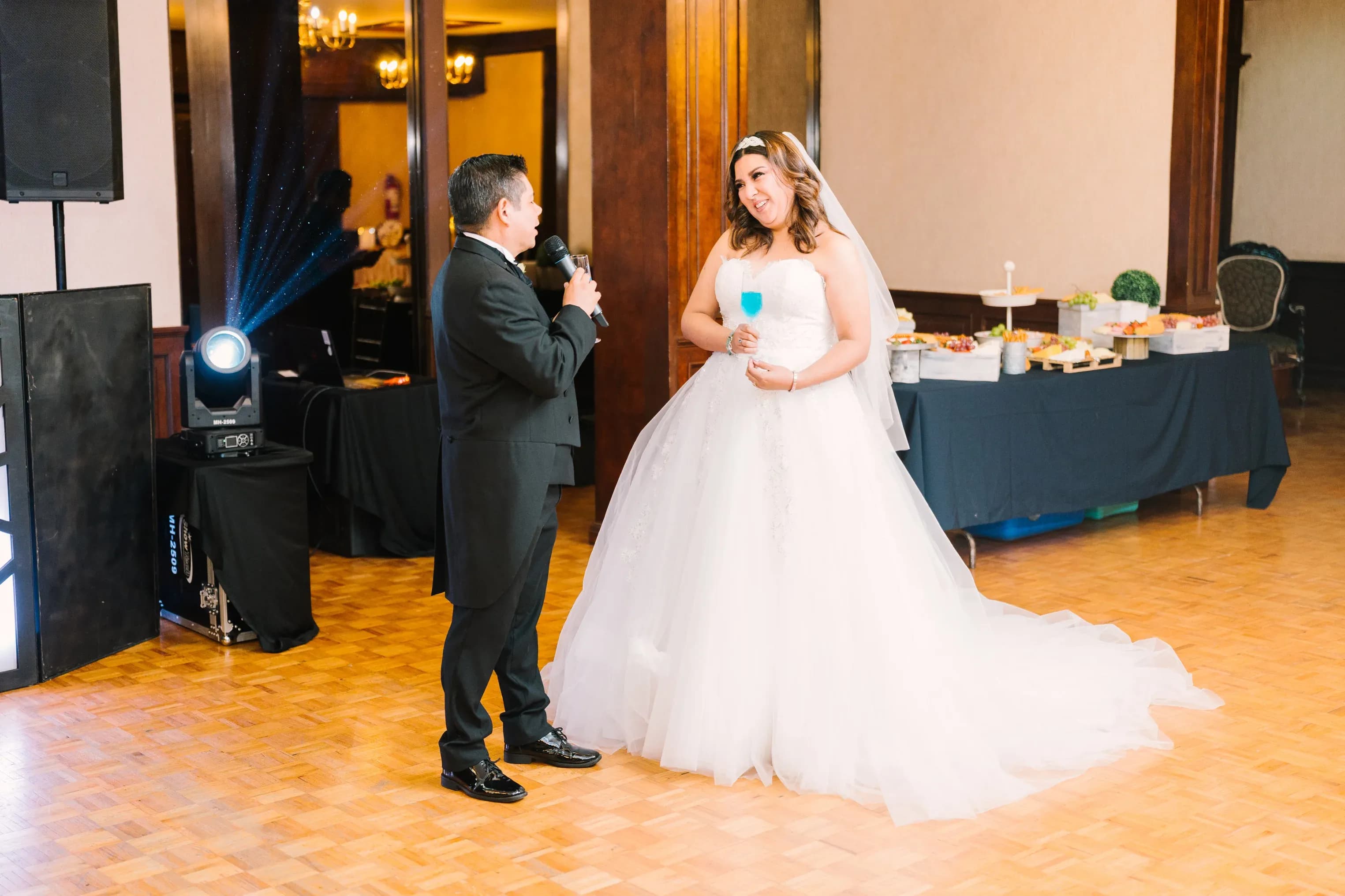 Novio con micrófono dando brindis junto a novia sonriente en pista de baile con mesa de bocadillos