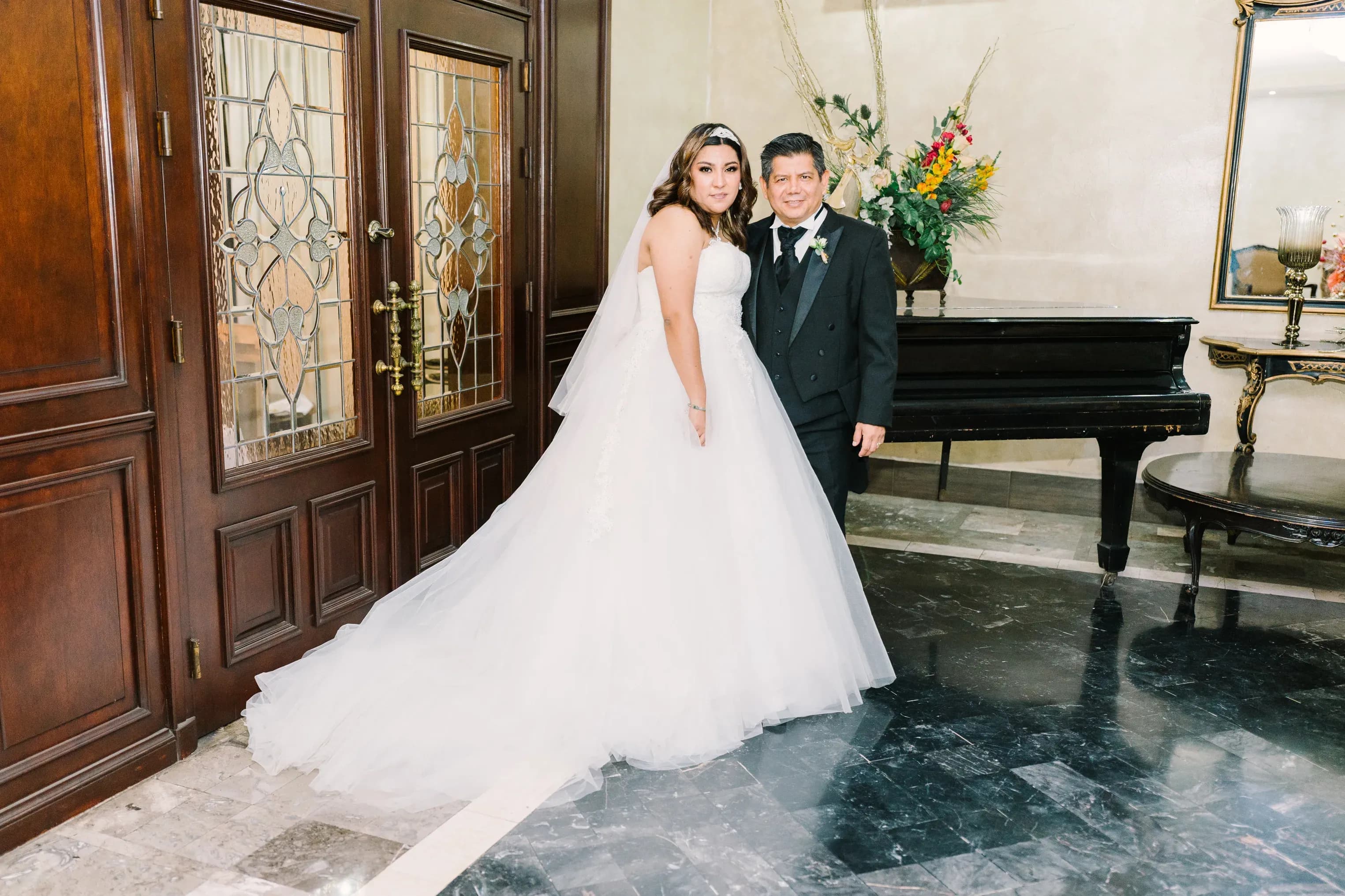 Pareja de novios posando en vestíbulo elegante con piano de cola y puerta de madera tallada