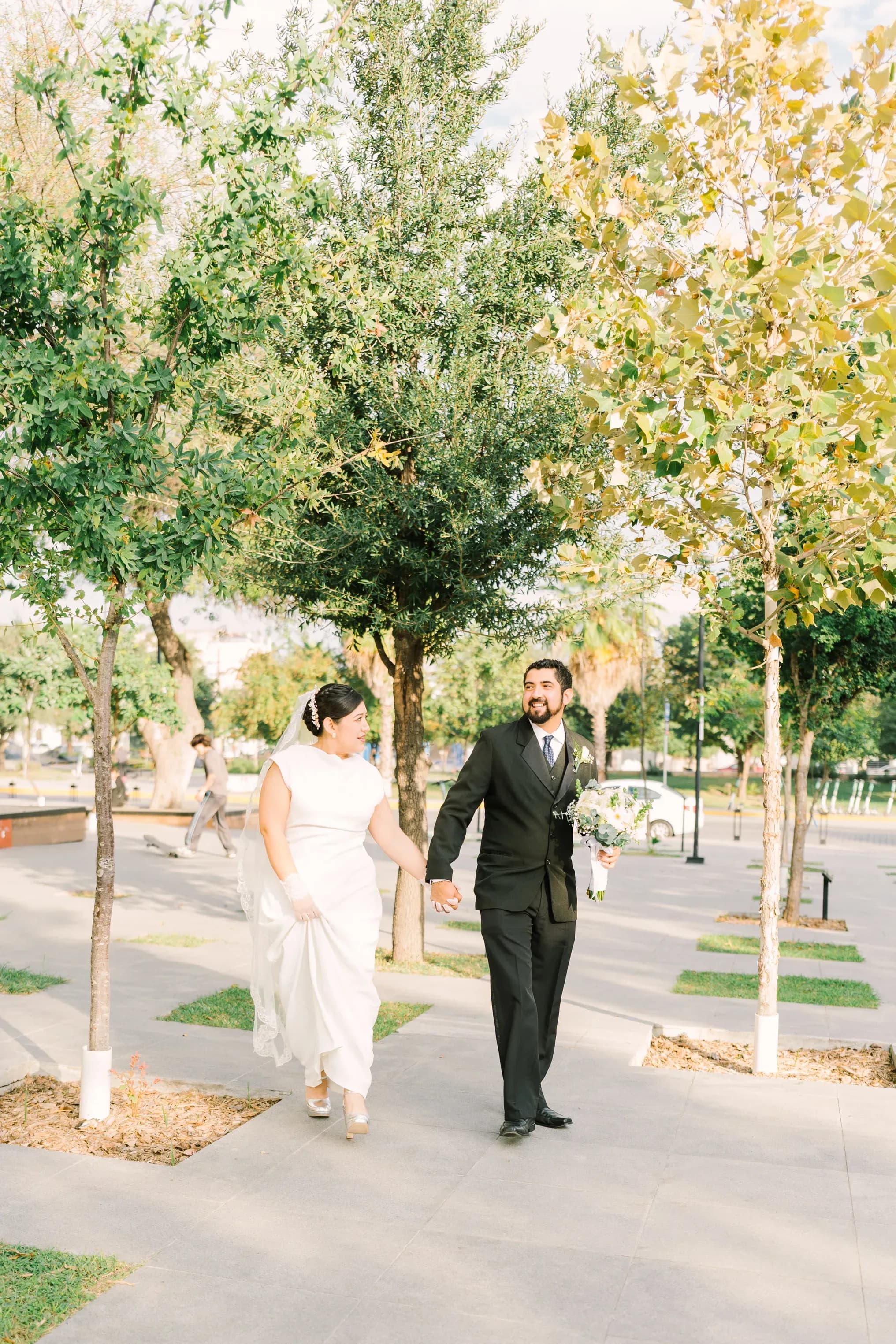 Novios tomados de la mano caminando entre árboles en plaza al aire libre durante sesión fotográfica