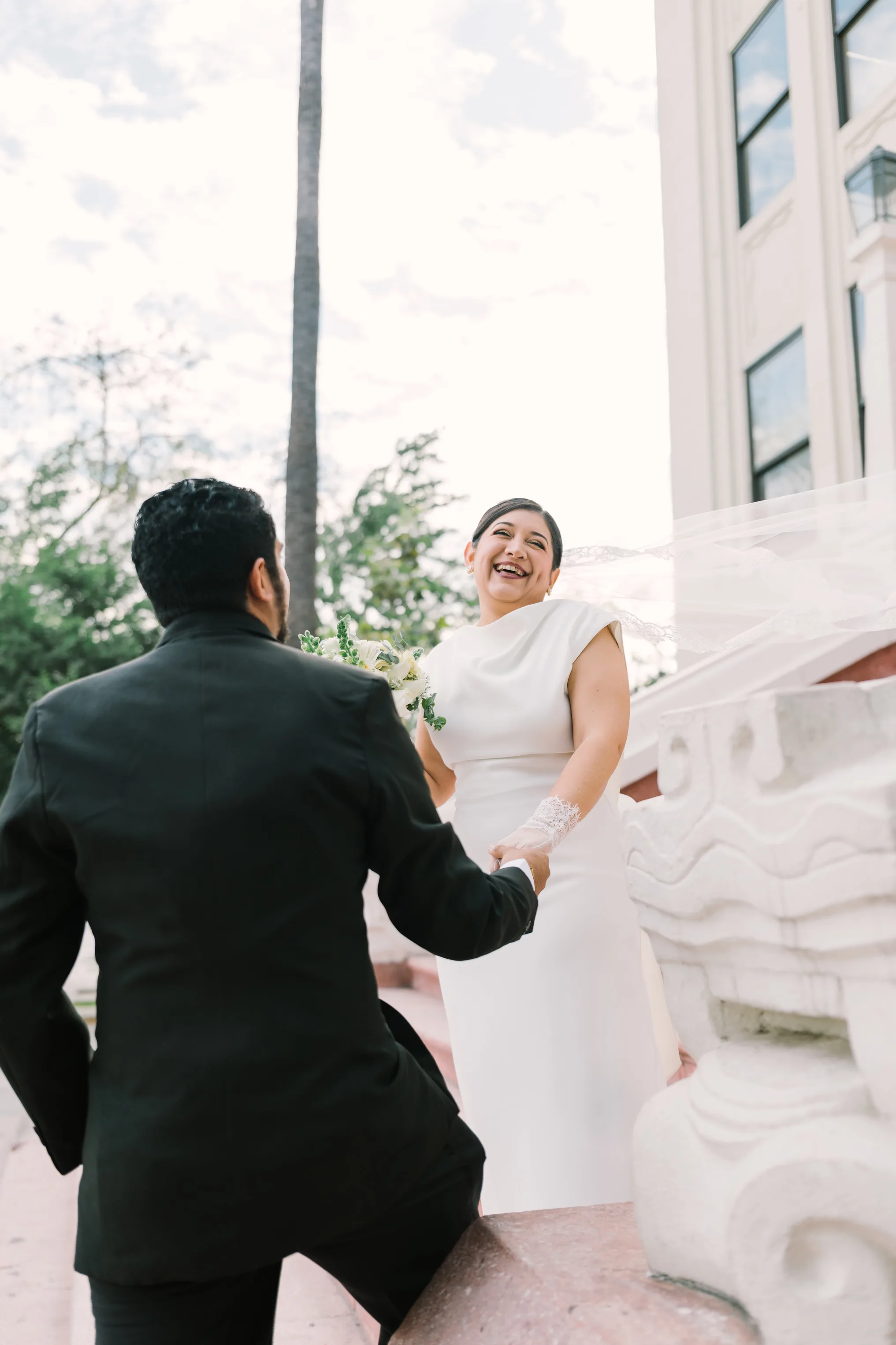 Novia sonriendo tomando la mano del novio en escalinata exterior durante sesión first look