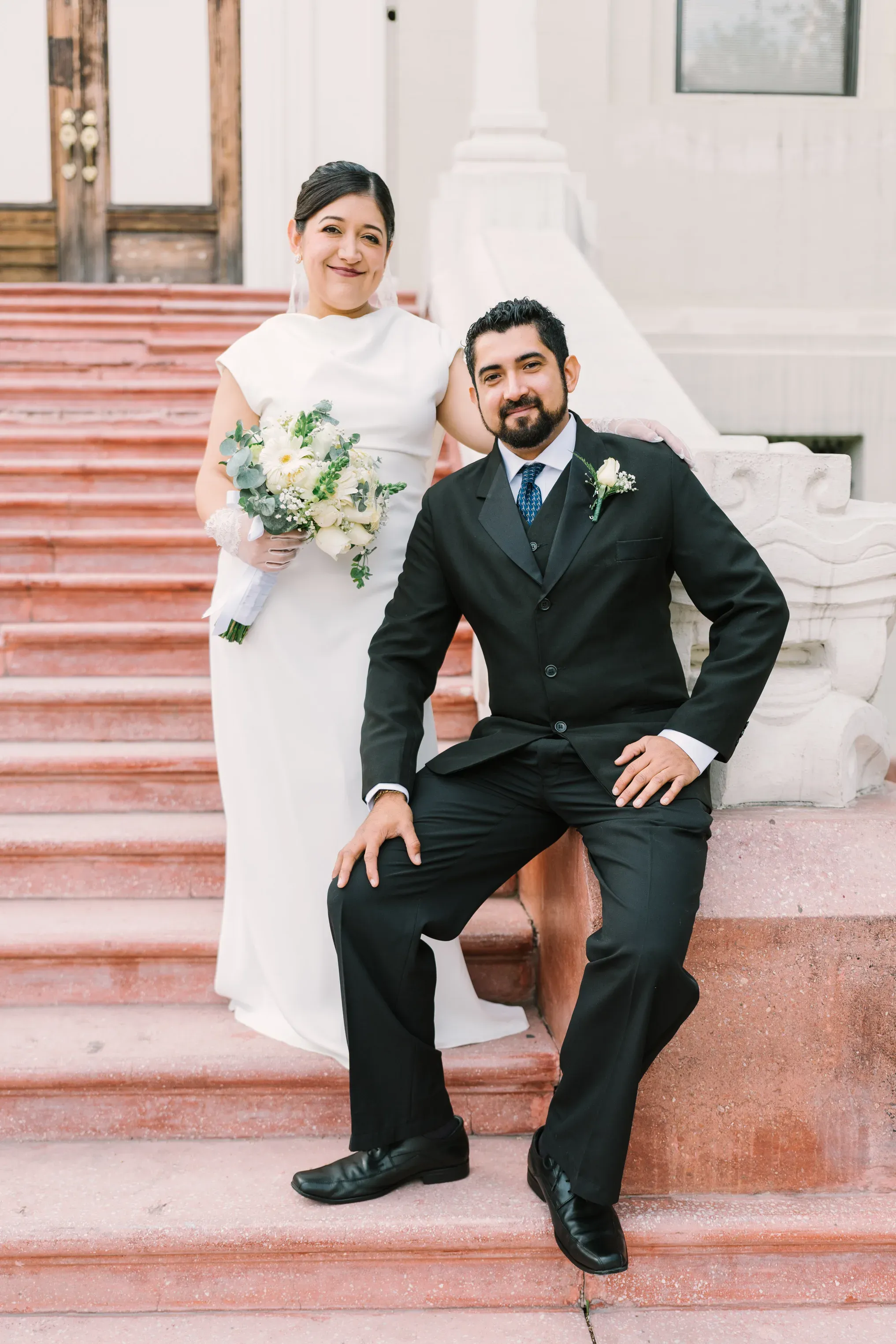 Novia de pie con ramo blanco y novio sentado en escaleras rojas de edificio histórico