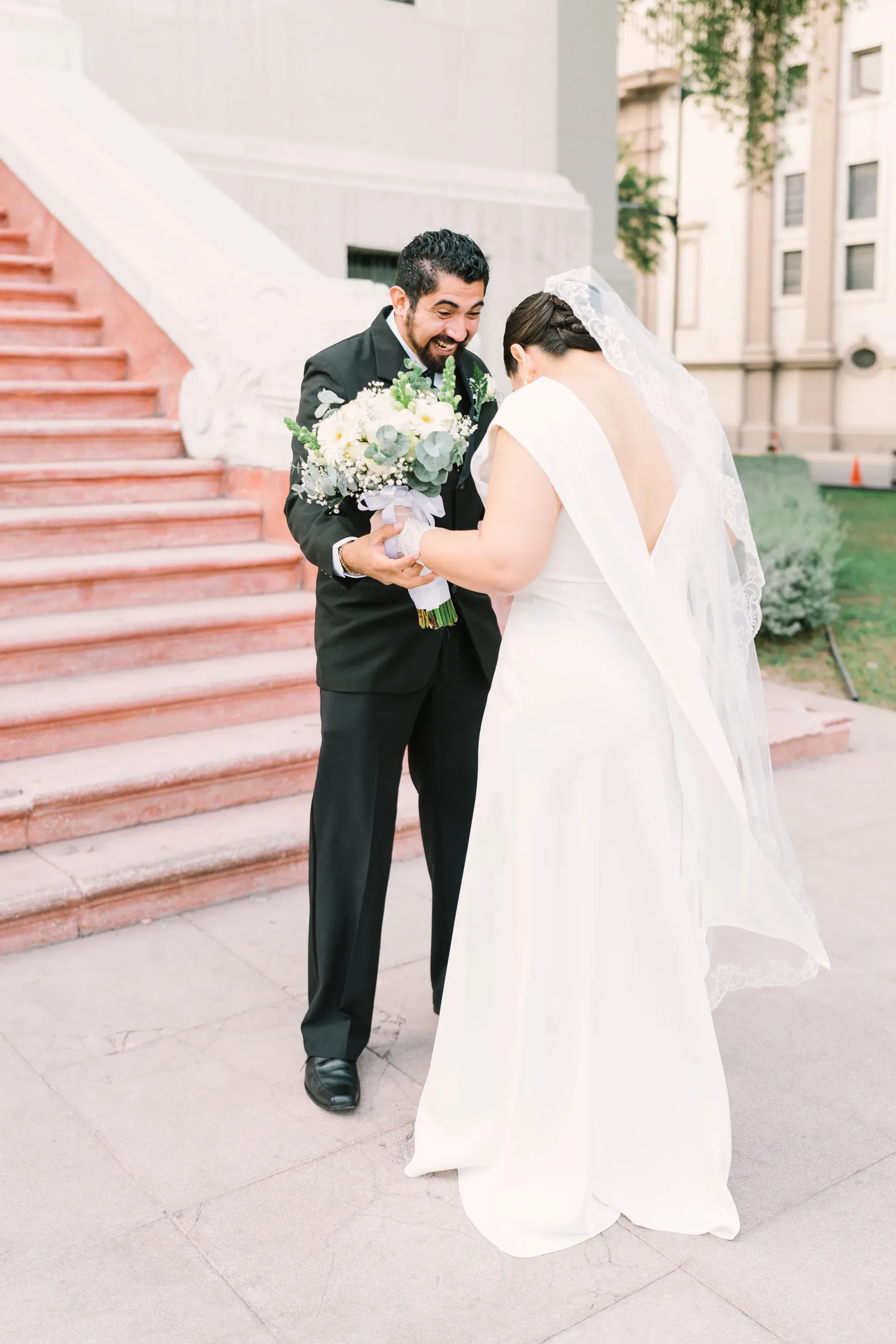 Novios riendo juntos revisando ramo de novia en escalinata exterior durante sesión de fotos