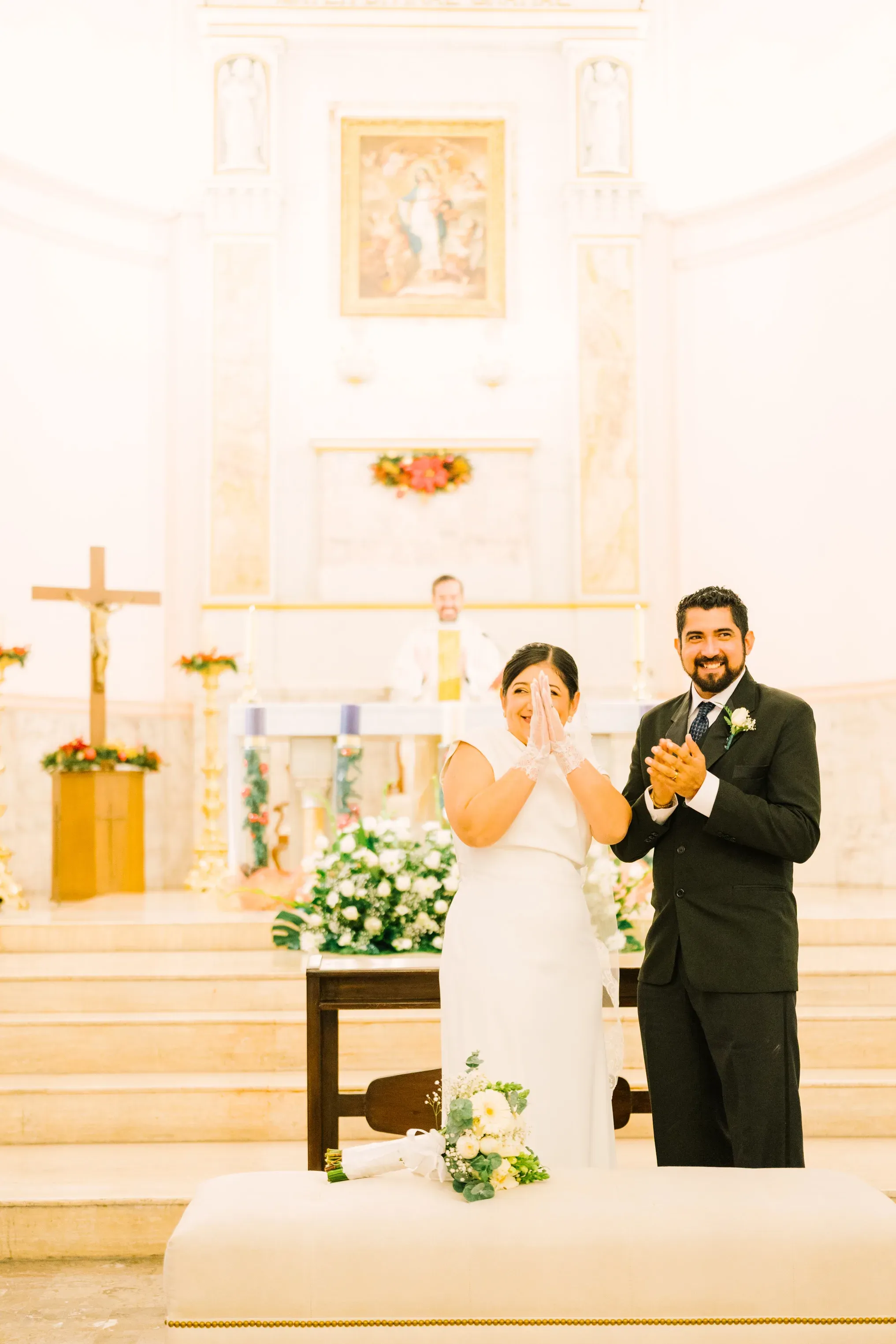 Pareja de recién casados aplaudiendo emocionados frente al altar de iglesia católica con sacerdote