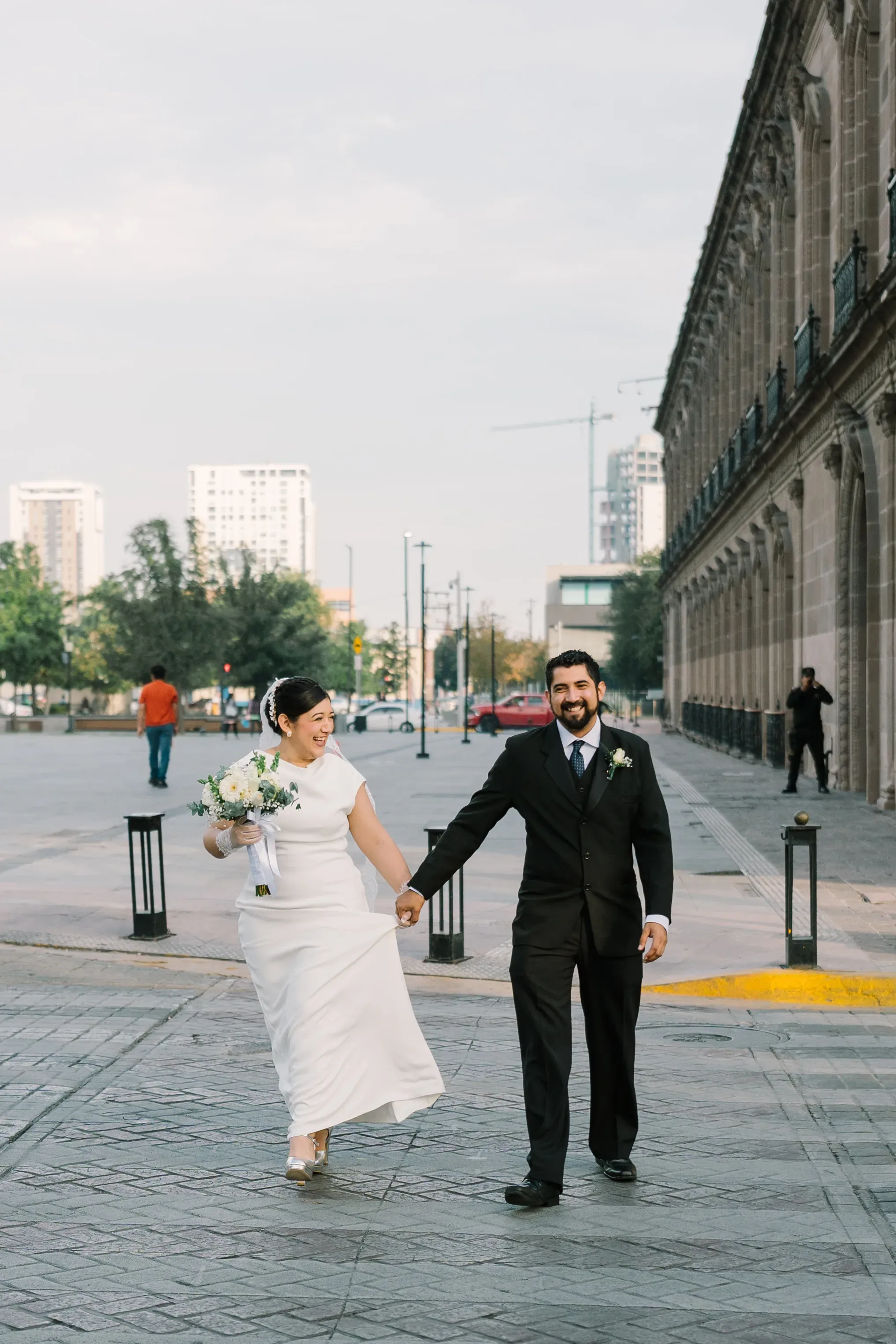 Novios tomados de la mano caminando felices frente a edificio neoclásico en Monterrey