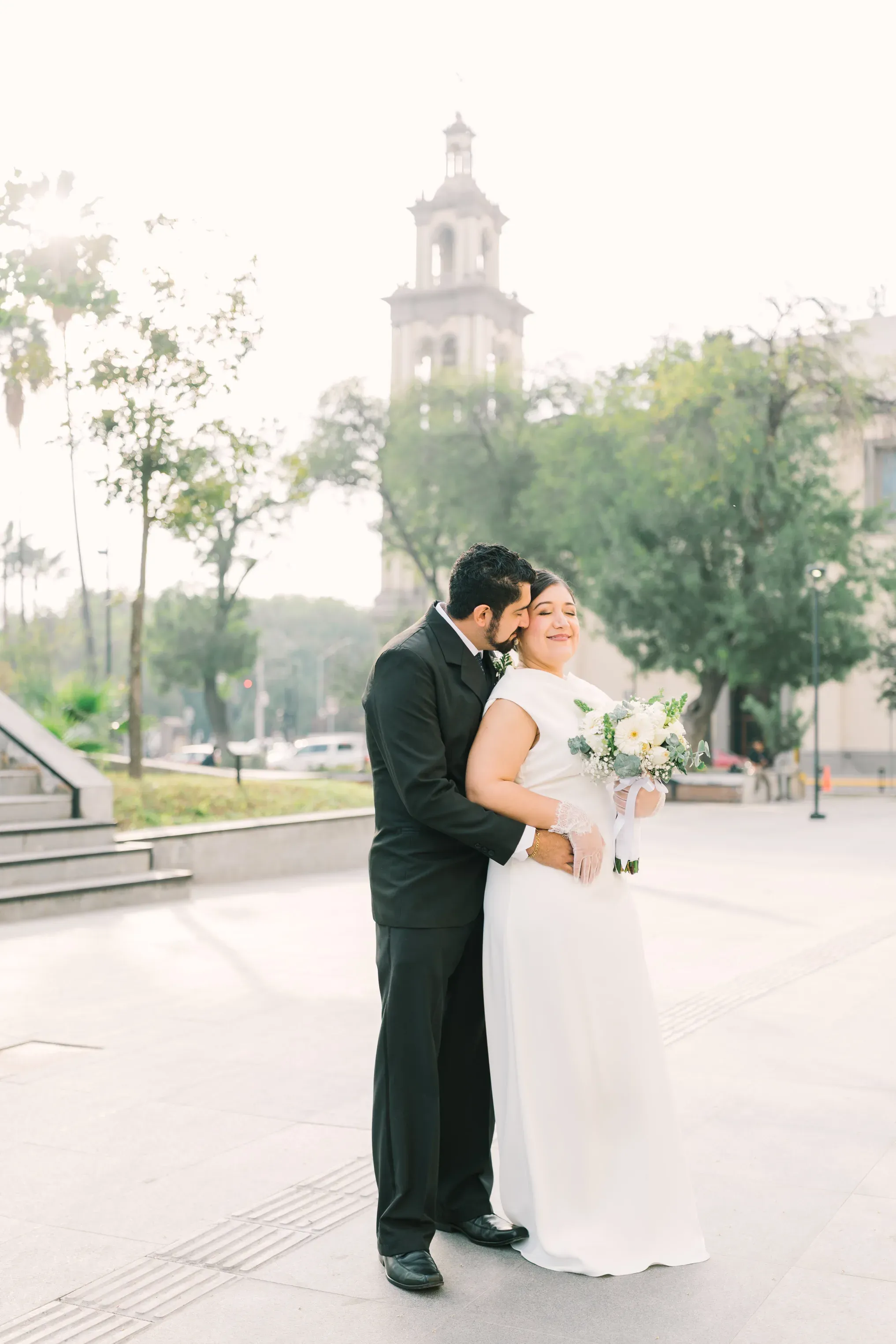 Novio abrazando y besando a novia sonriente con torre de iglesia al fondo en plaza de Monterrey