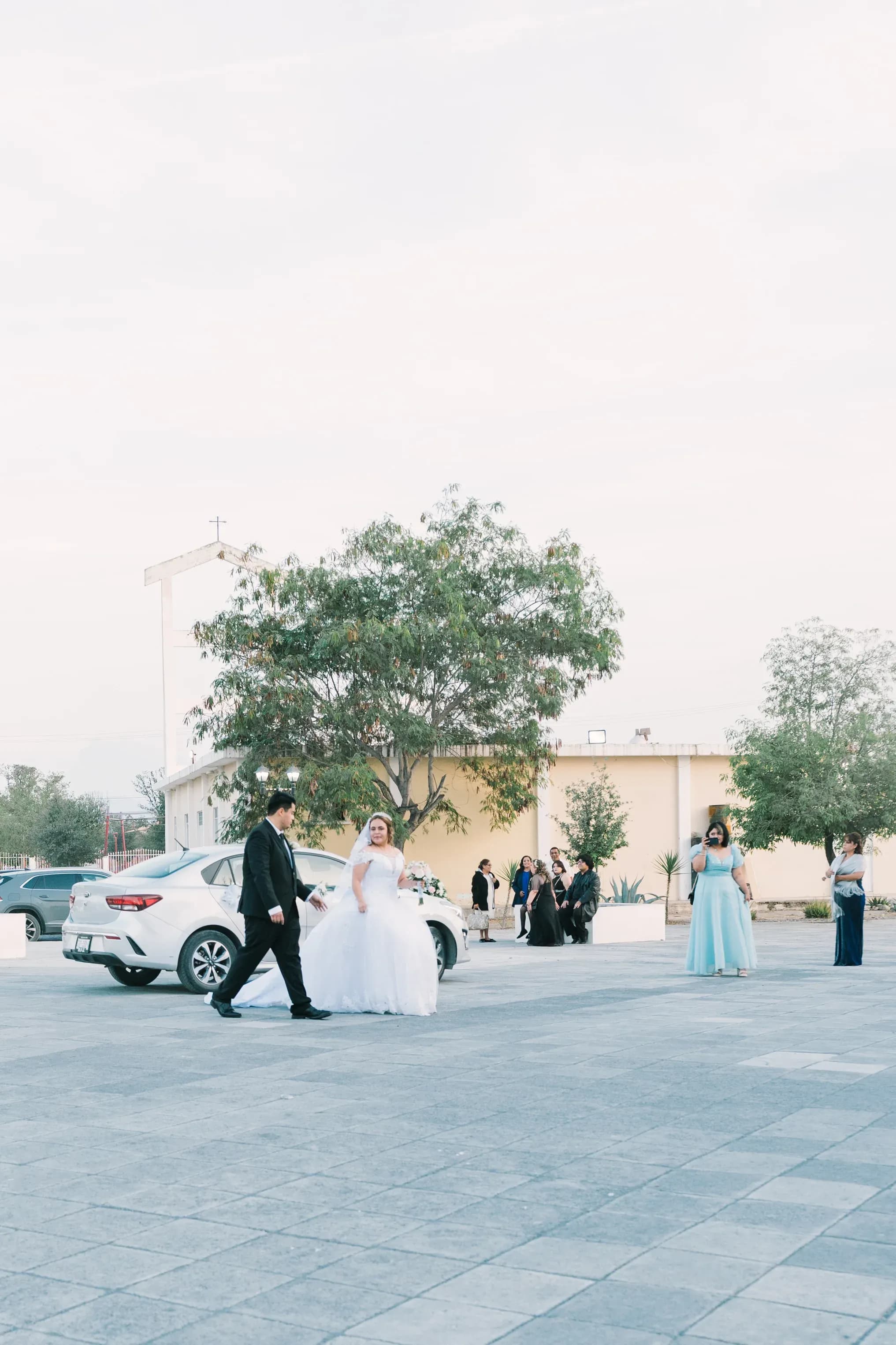 Novios llegando a iglesia en estacionamiento con dama de honor y familiares observando
