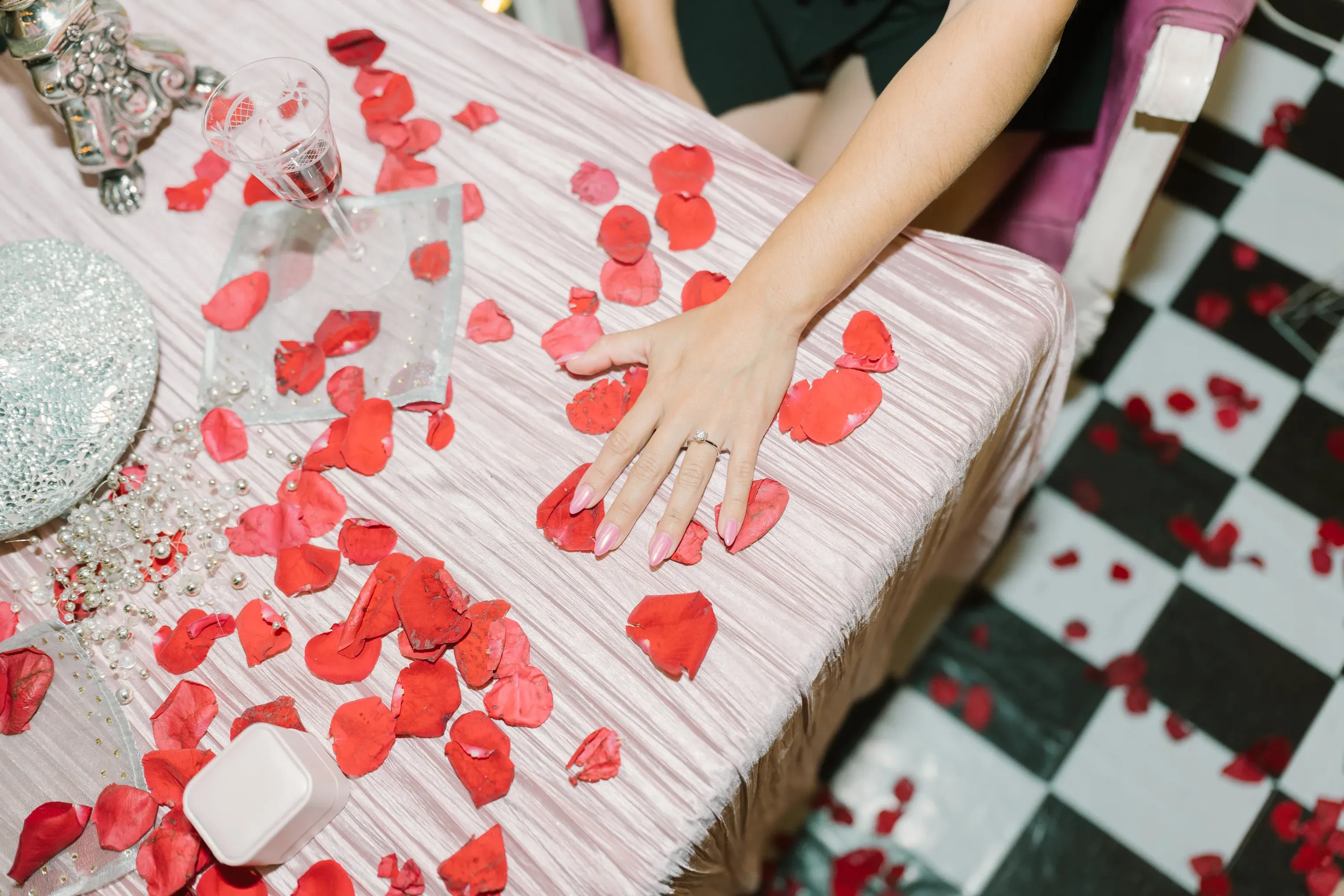 Mano de mujer con anillo de compromiso y uñas rosas sobre mesa decorada con pétalos de rosa rojos