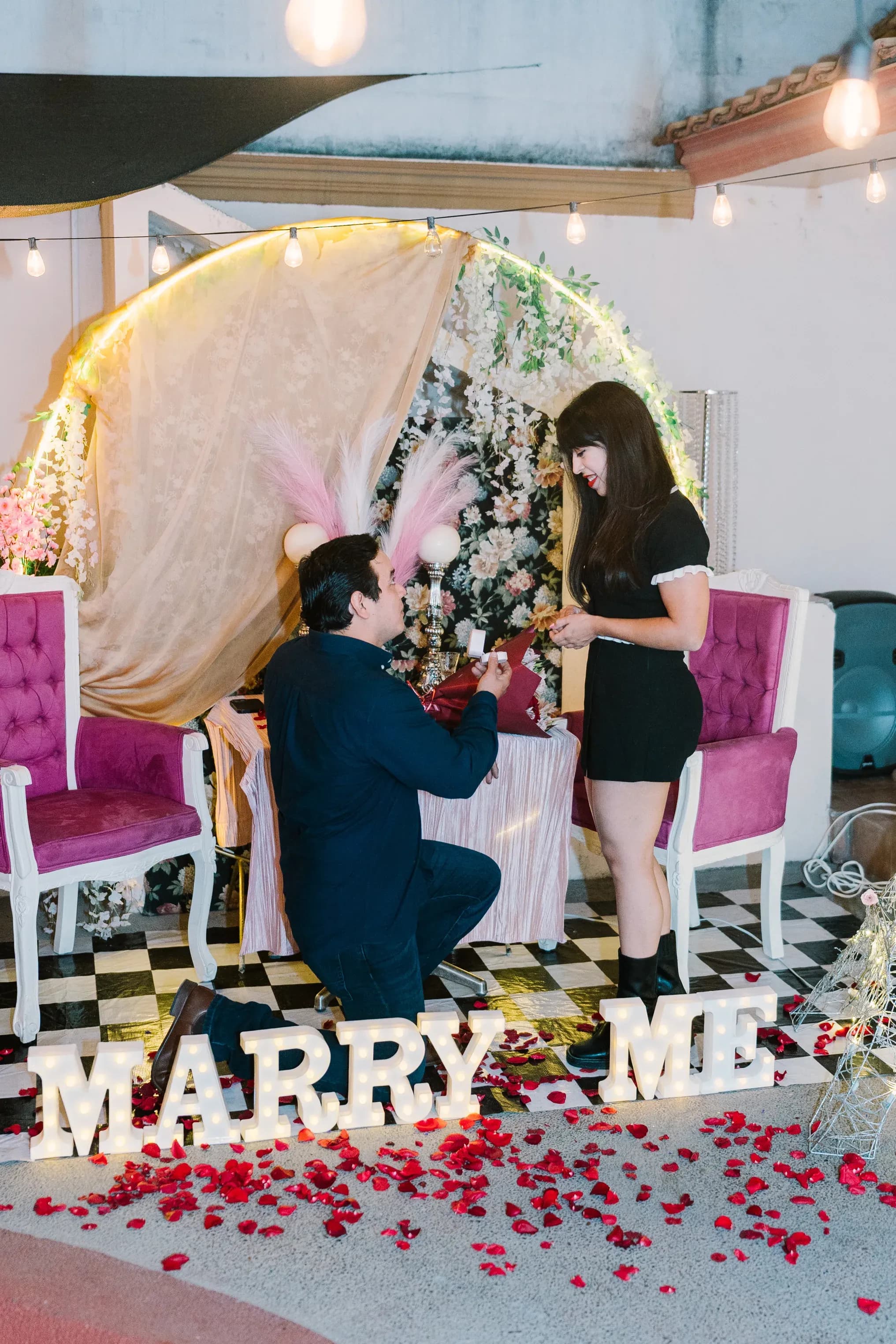 Hombre arrodillado pidiendo matrimonio con letrero MARRY ME iluminado y pétalos de rosa rojos