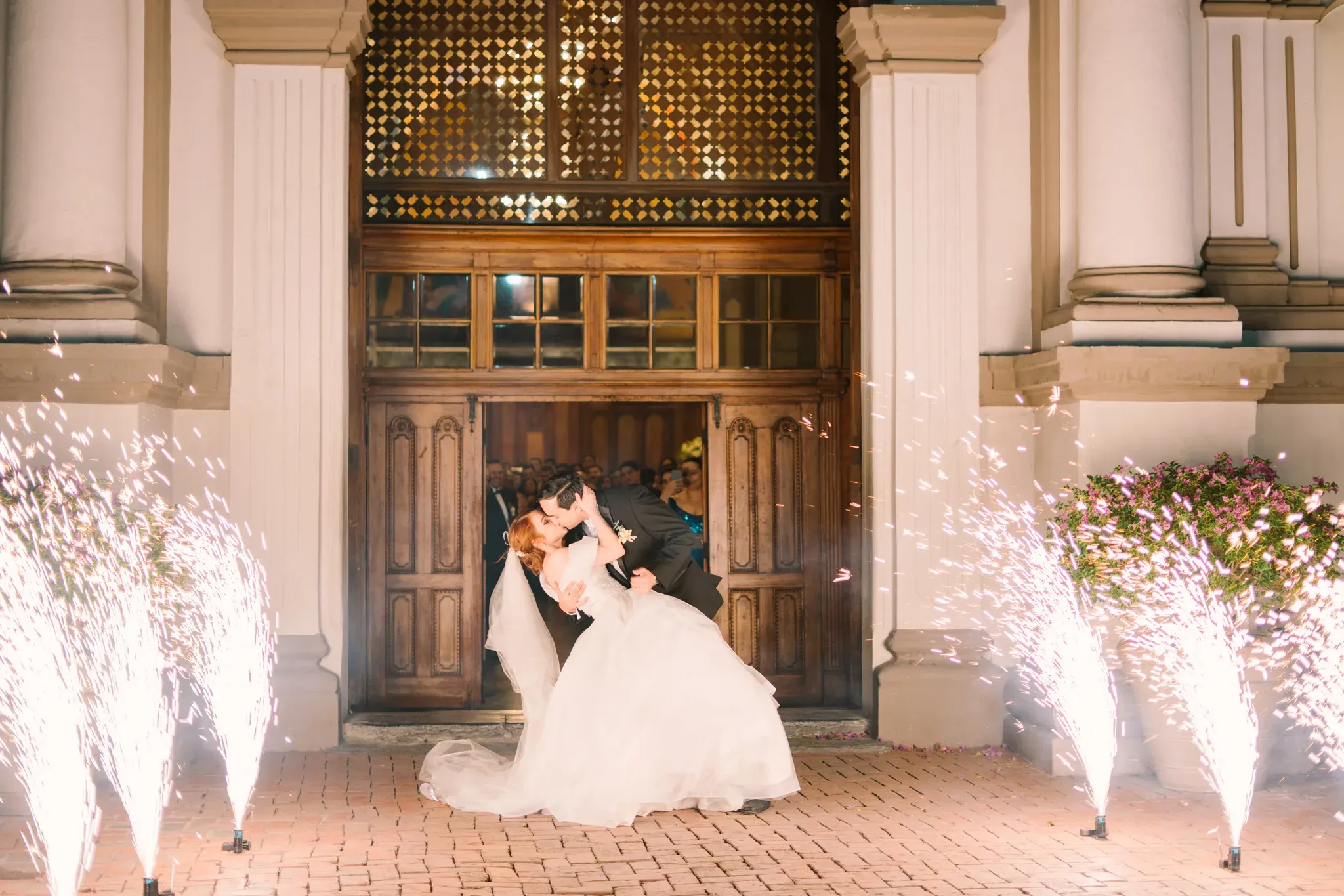 Novio cargando a novia en dip kiss a la salida de iglesia con fuentes de chispas en la noche
