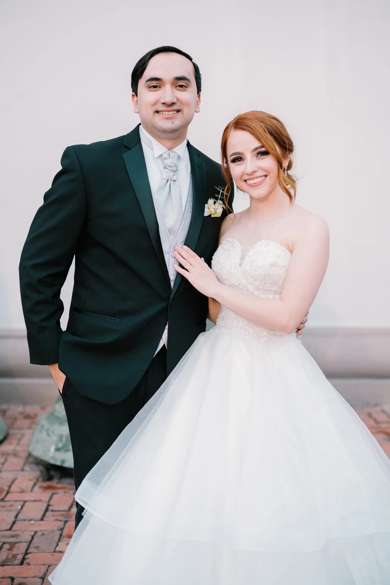 Pareja de novios posando juntos frente a pared blanca, novia con vestido de princesa y pelo rojo