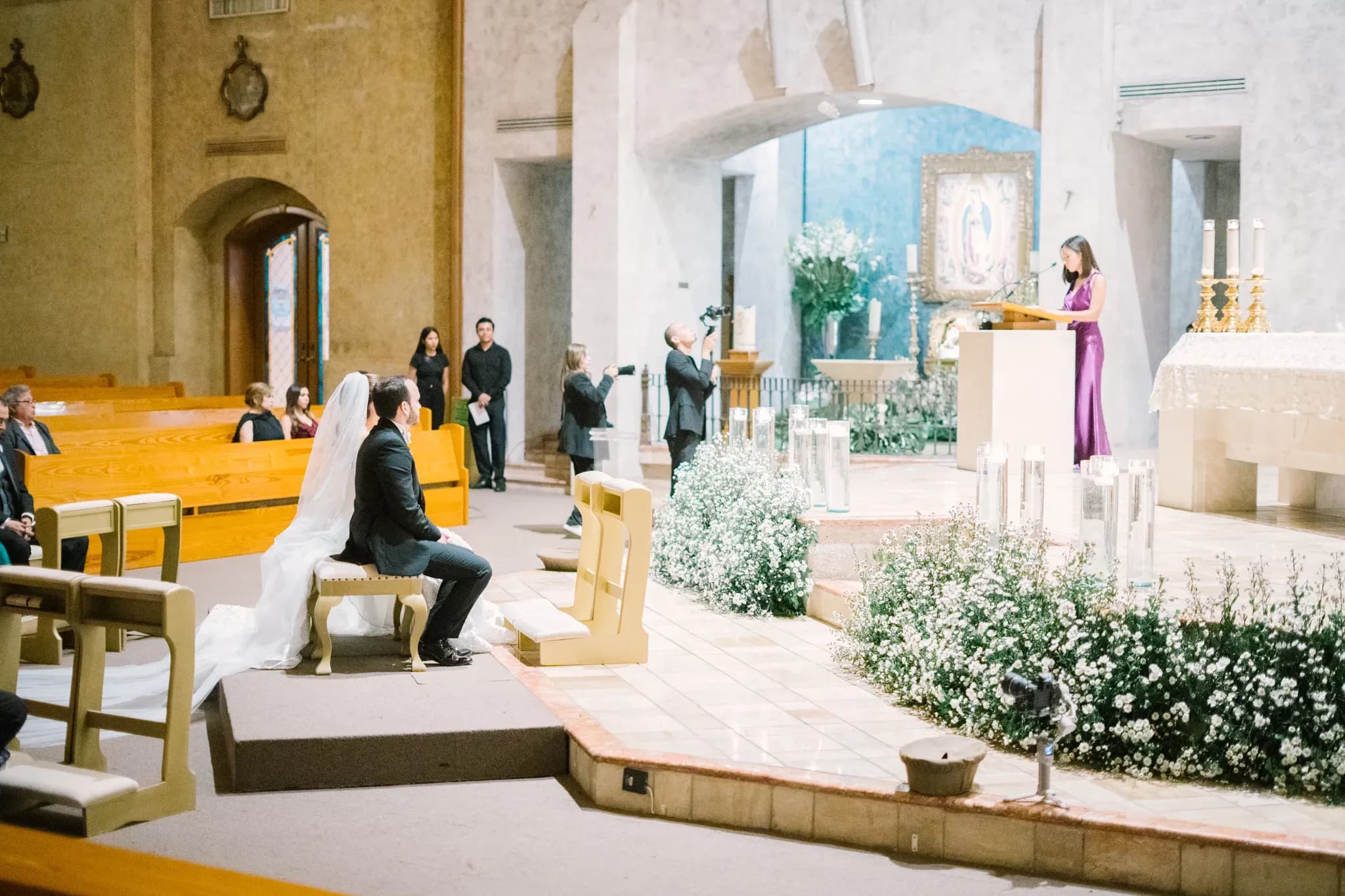 Novia y novio sentados en reclinatorio durante ceremonia religiosa con lectora en púlpito de iglesia