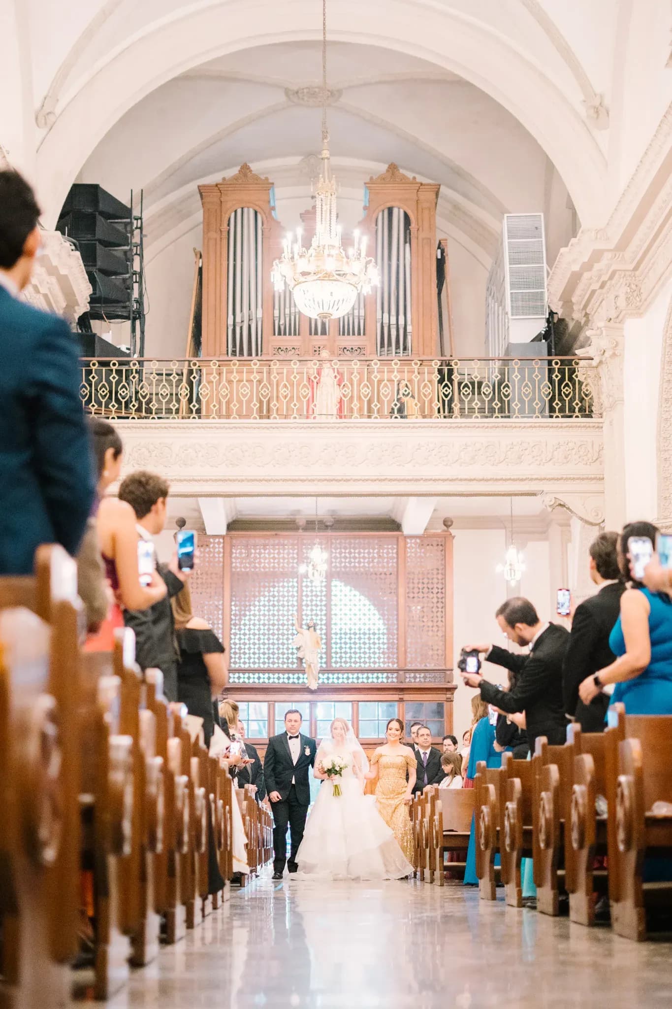 Novia entrando al altar acompañada por sus padres en iglesia con órgano y candelabro de cristal