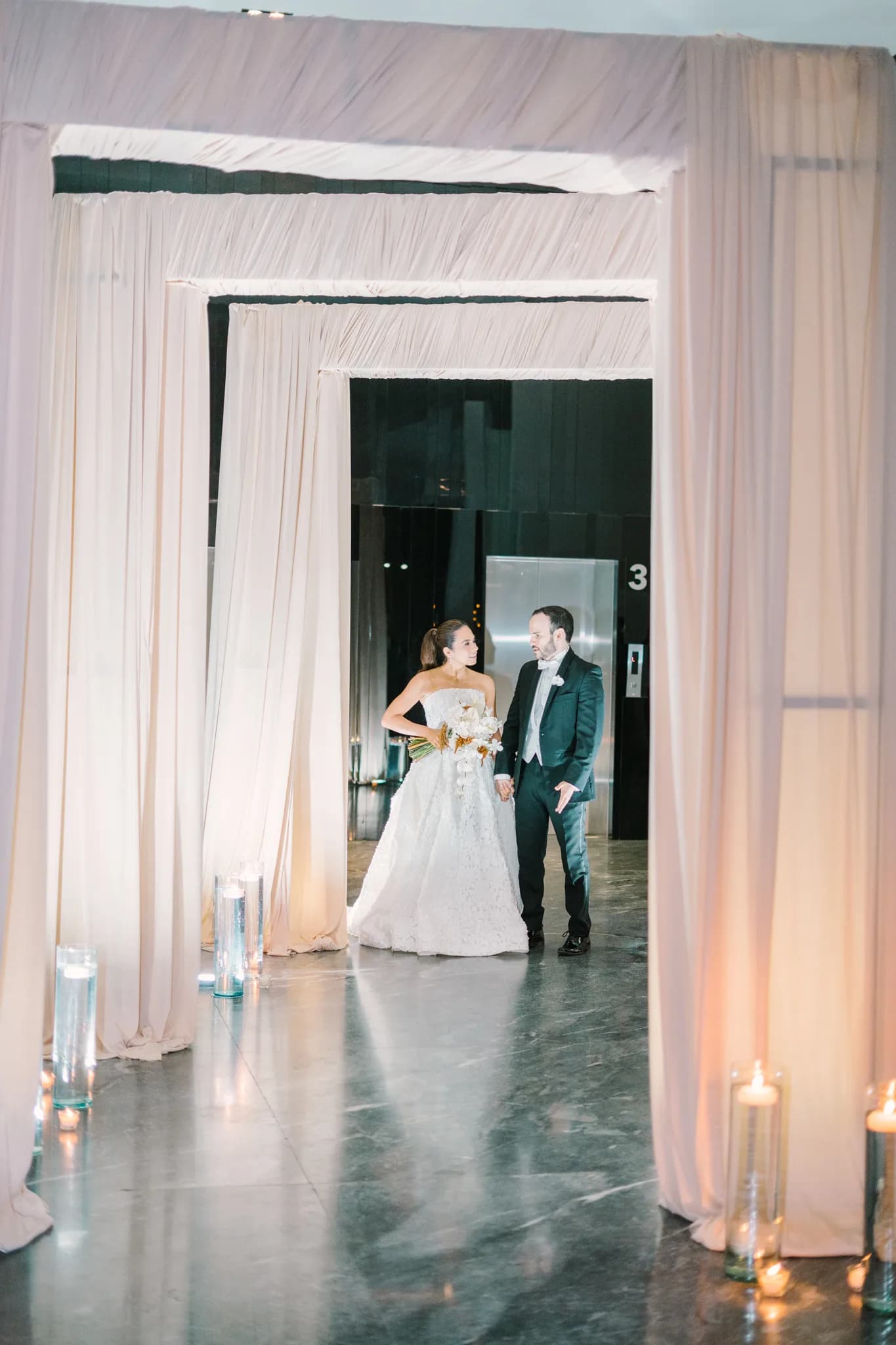Pareja de recién casados entrando a salón de recepción por pasillo de cortinas blancas con velas
