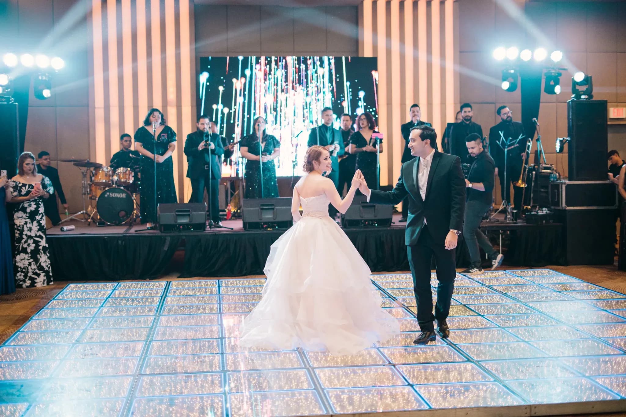Novios bailando vals en pista iluminada con banda en vivo y pantalla LED en recepción de boda
