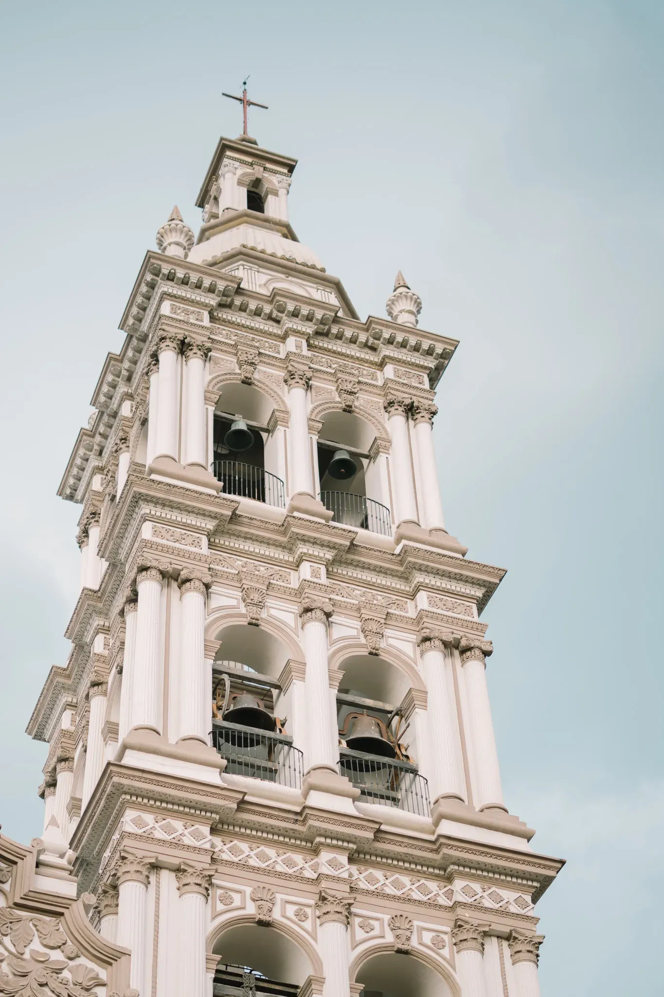 Torre campanario barroca blanca de iglesia con campanas y cruz en lo alto contra cielo nublado