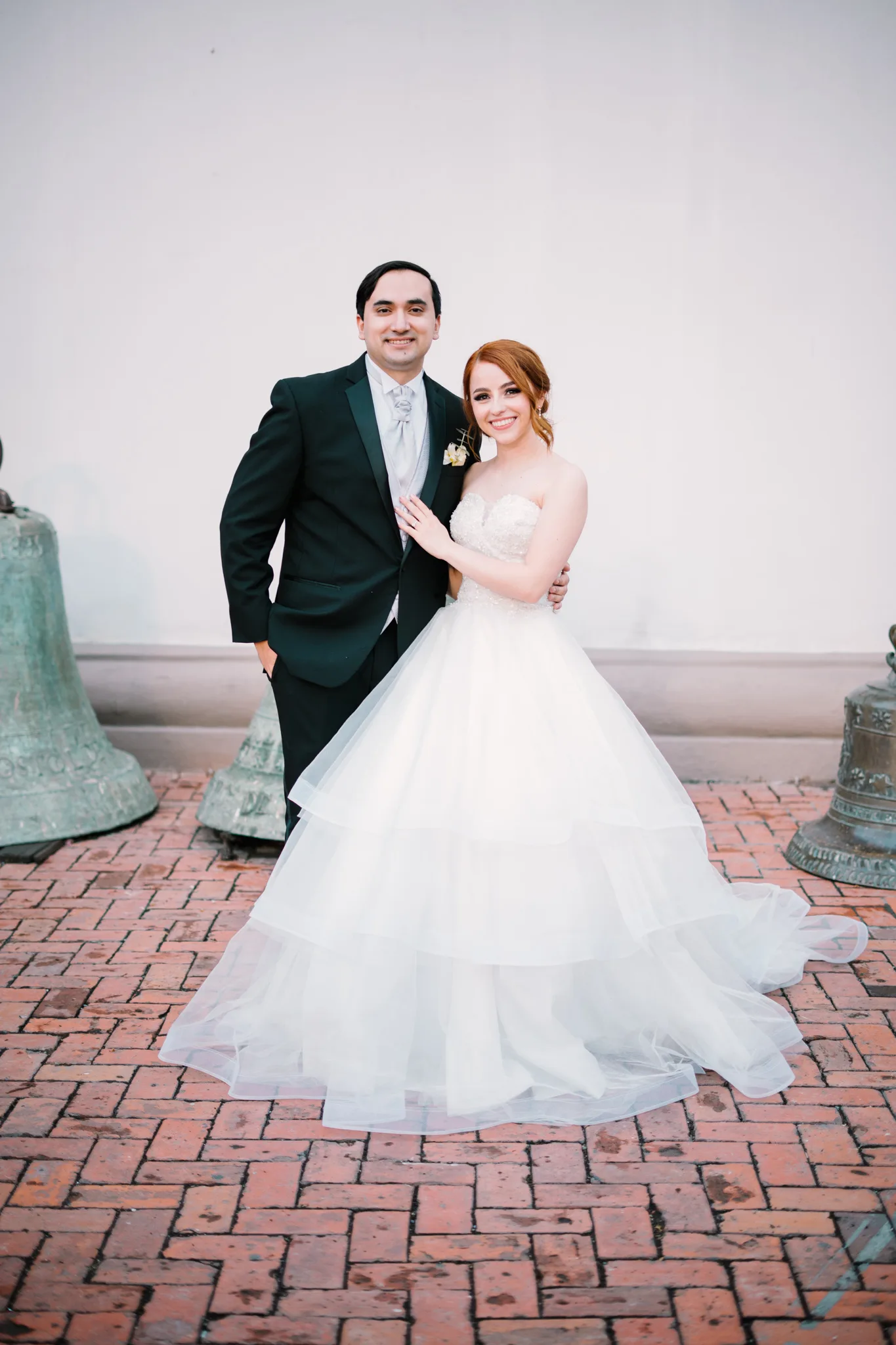 Novios sonriendo a cámara junto a campanas históricas de iglesia en terraza de ladrillo