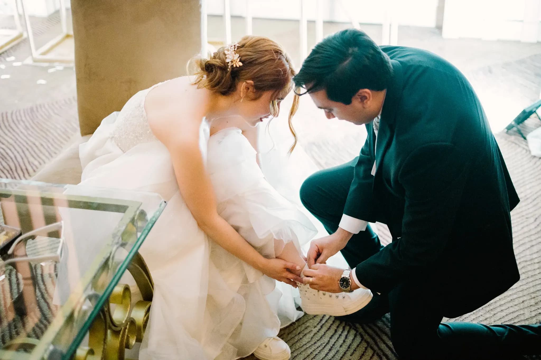 Novio arrodillado colocando tenis blancos a la novia durante la recepción de boda
