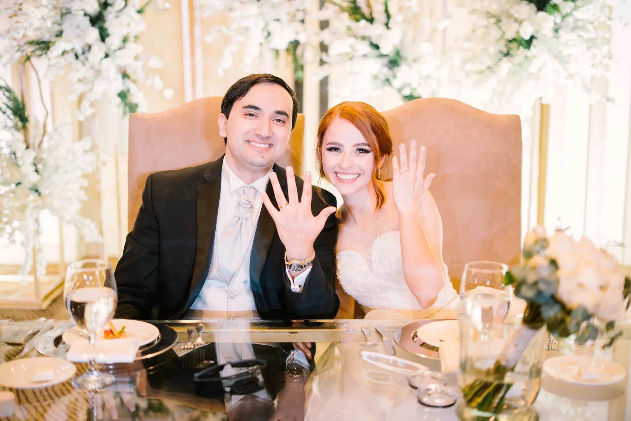 Recién casados mostrando sus anillos de bodas sonriendo en mesa de honor con flores blancas